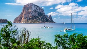 View of Ibiza with a rocky island in the background and sail boats floating on the sea in front.